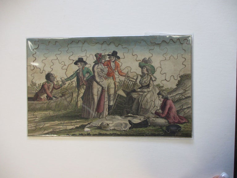 Item #38153 57 PIECE JIGSAW PUZZLE, LATE 18TH CENTURY, DEPICTING A PICNIC SCENE OF FIVE ARISTOCRATIC CHARACTERS PREPARING TO PICNIC ON SHORE. A BLACK SERVANT OR SLAVE IS IN THE ROWBOAT WHICH IS TIED TO THE TREE ON SHORE; HE IS HANDING A BOTTLE TO ONE OF THE MEN. ONE MAN IS SEATED ON THE GROUND WHILE HIS LADY IS SEATED ON A CHAIR BY HIS SIDE. A SECOND MAN IS SETTING UP A CHAIR FOR HIS LADY. Jigsaw Puzzle.